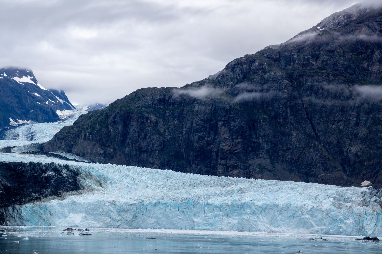 Towering glacier face glows blue against dark mountain cliffs in misty fjord.