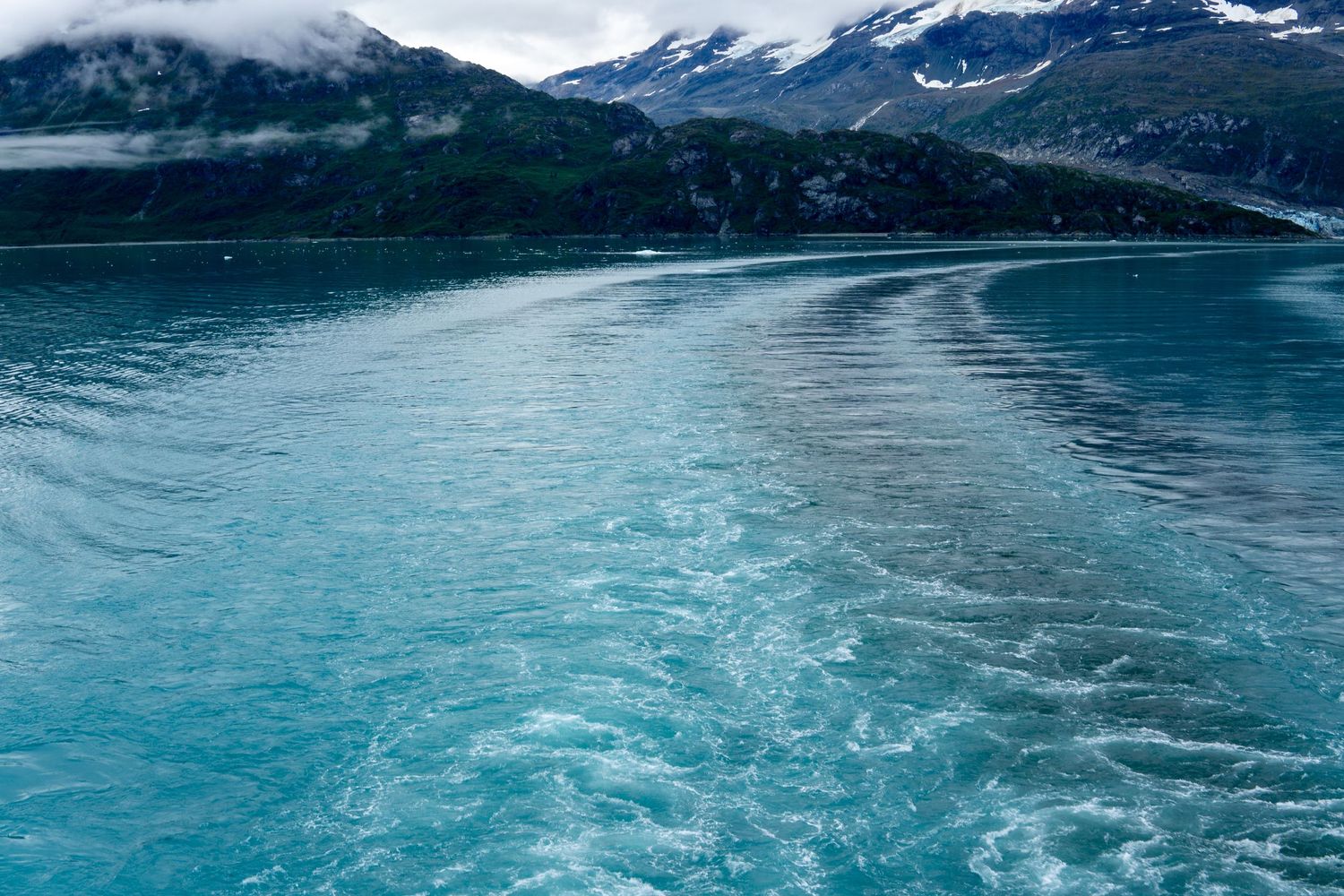 Turquoise wake patterns ripple through glacial waters with snow-capped mountains in background.