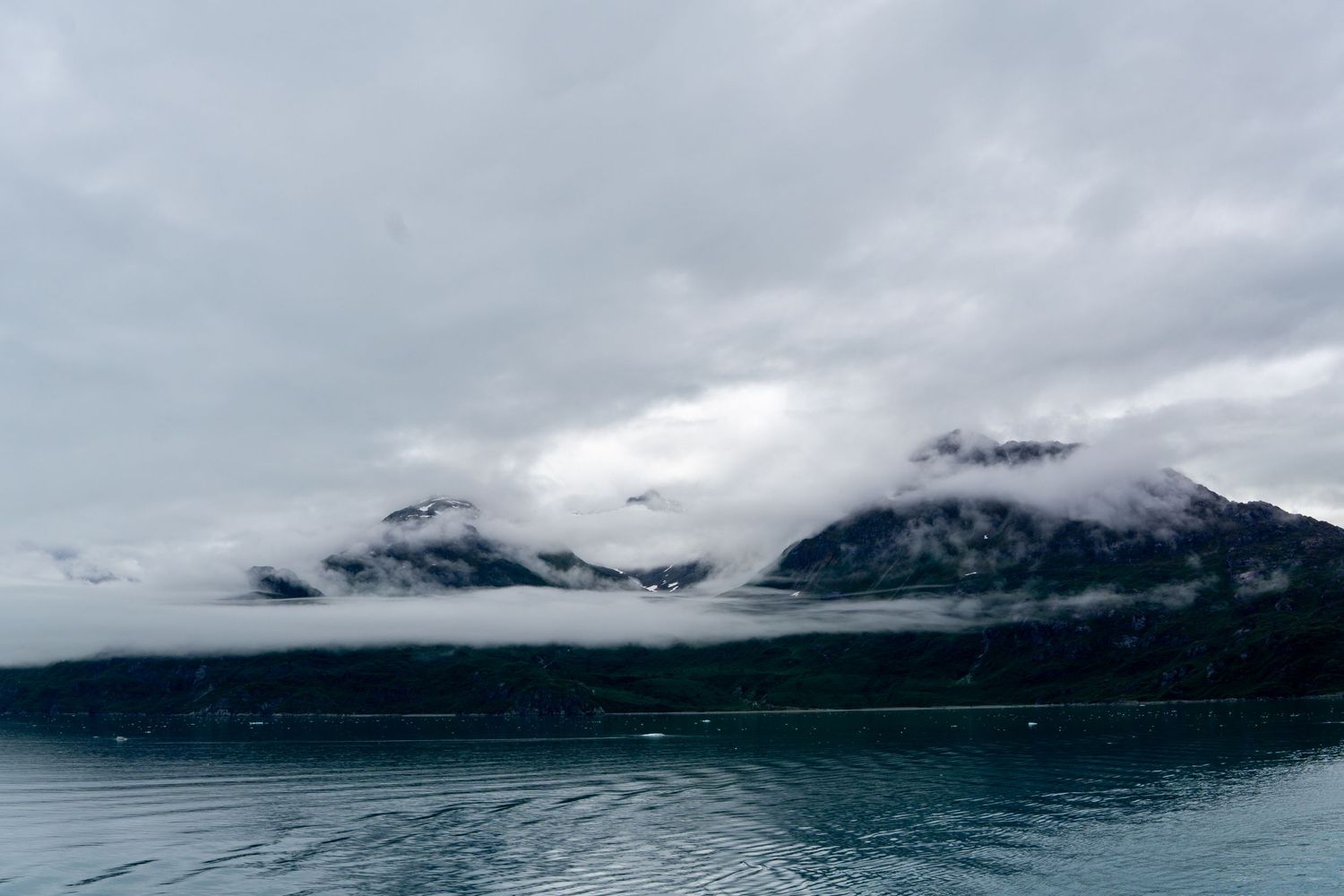 Misty mountains rise above calm waters with low-hanging clouds creating a moody atmosphere.