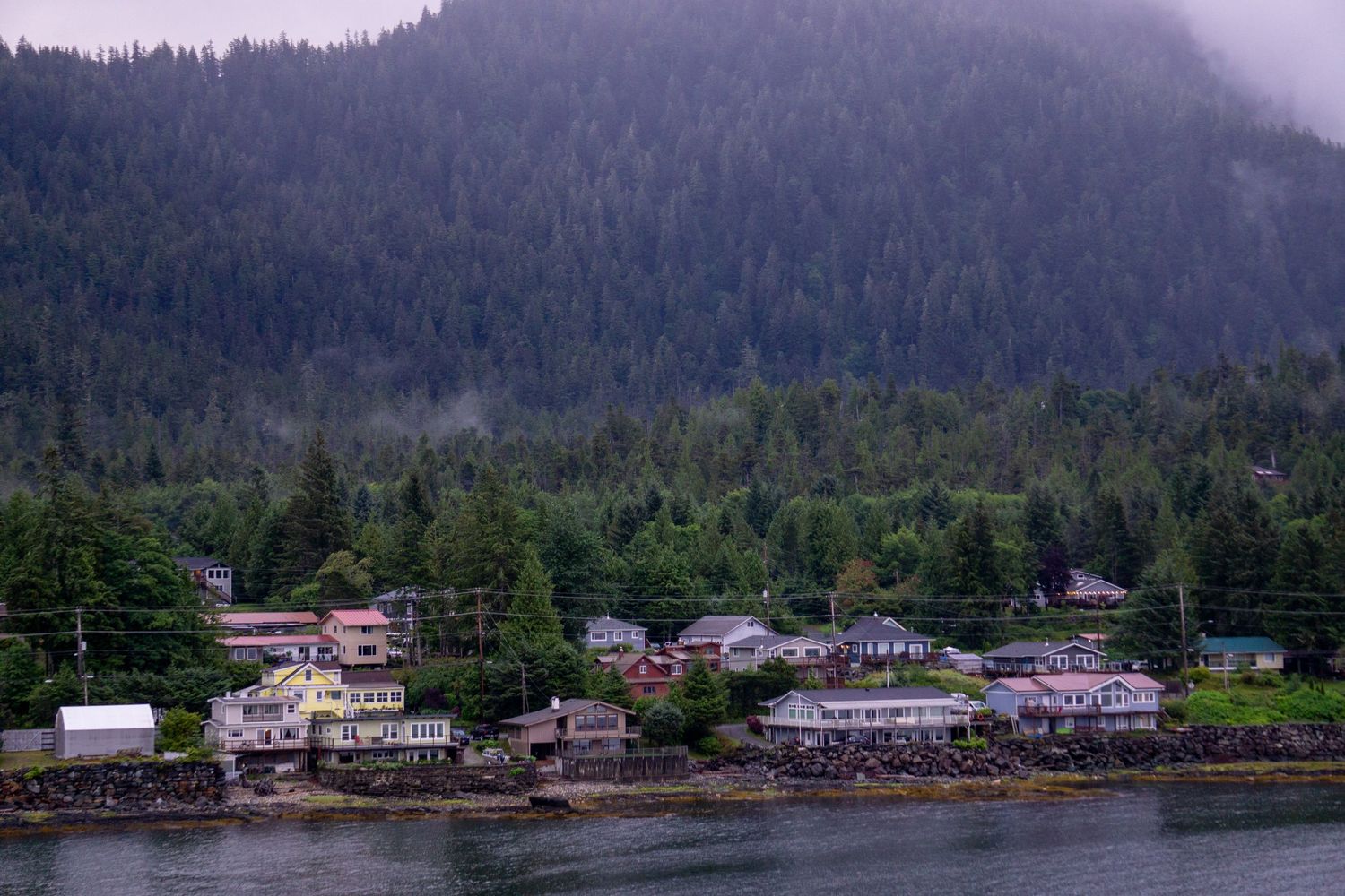 Waterfront homes and buildings nestled along a foggy coastal shoreline with forested mountains in the background.