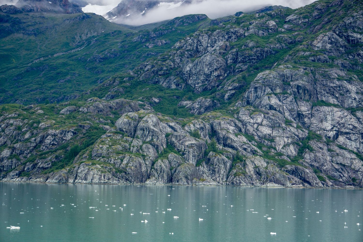 Steep rocky cliffs rise from turquoise glacial waters with low-hanging clouds in an Alaskan fjord.