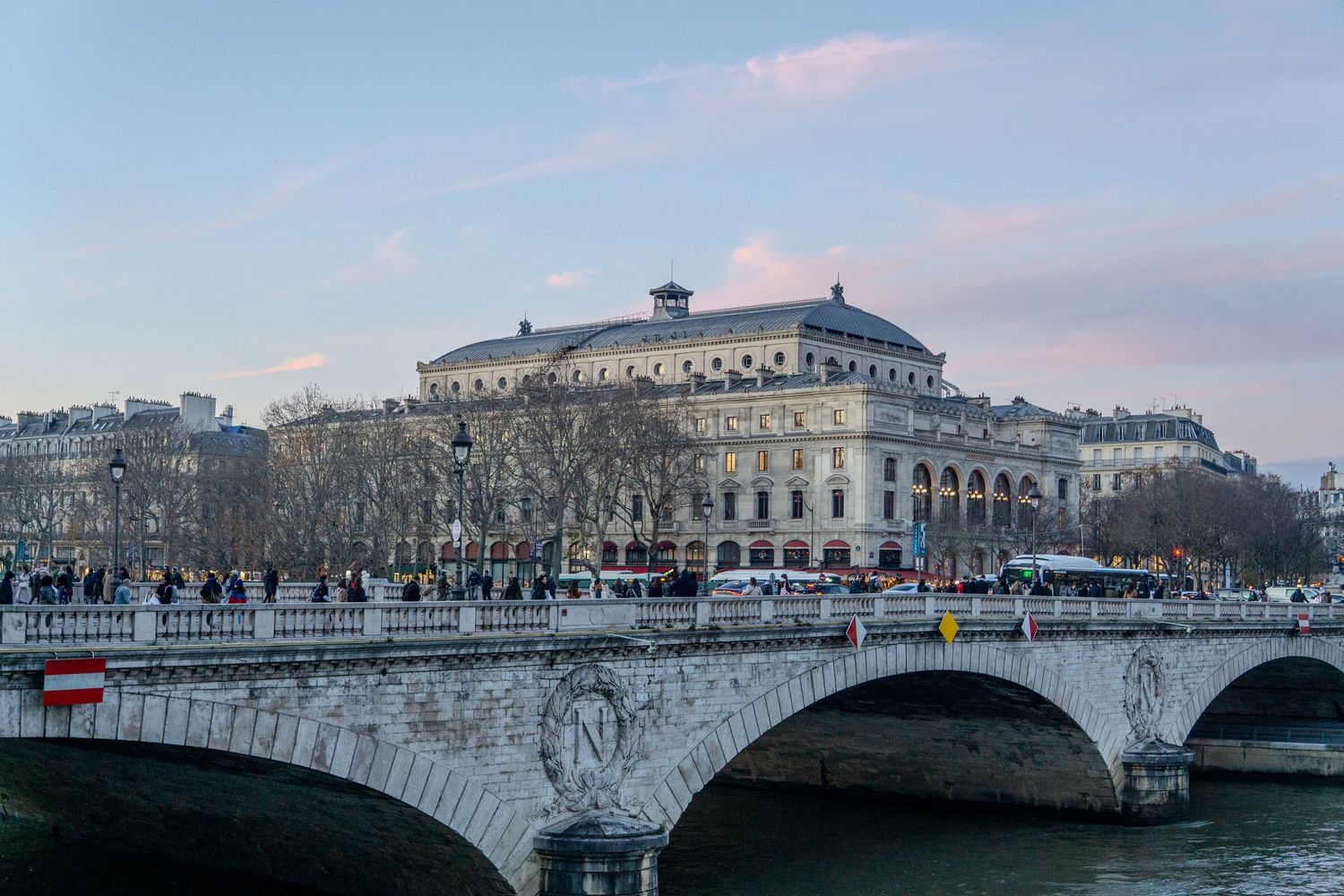 Historic stone bridge spans the Seine River at dusk with the illuminated Theatre du Chatelet building in Paris, France.
