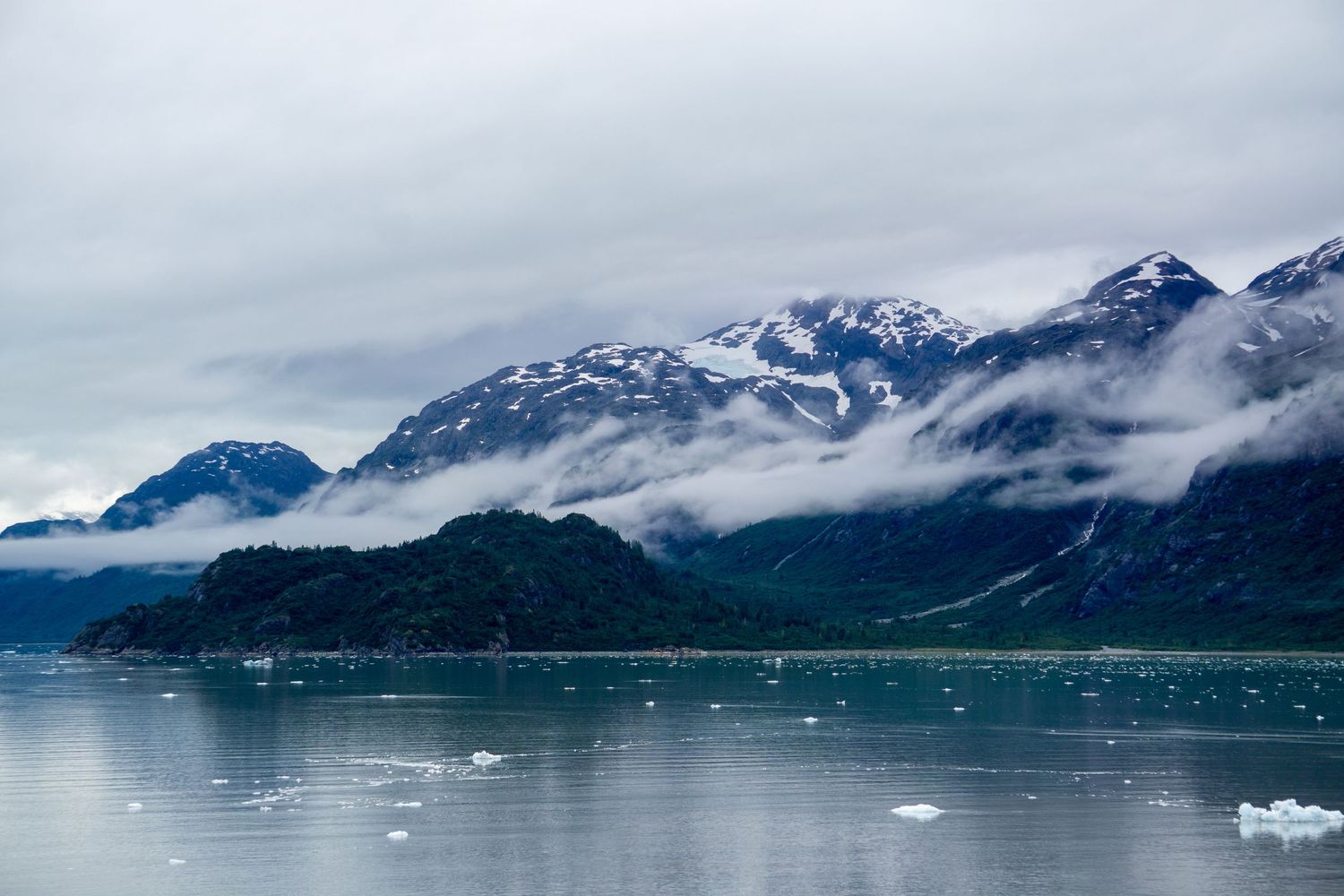 Fog-shrouded mountains with snow caps rise above icy waters dotted with floating ice chunks.
