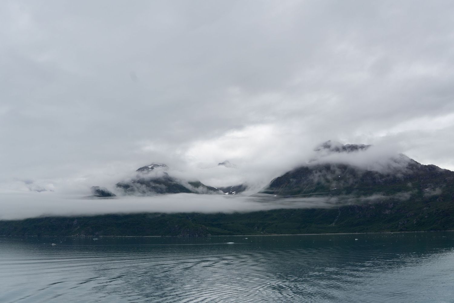 Fog-shrouded mountain peaks loom over dark waters in dramatic Alaska scenery.
