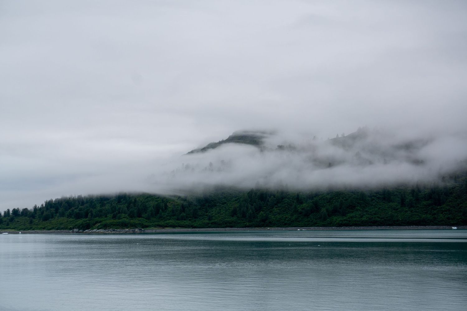 Misty clouds hover over a forested mountain slope reflecting in still waters below.