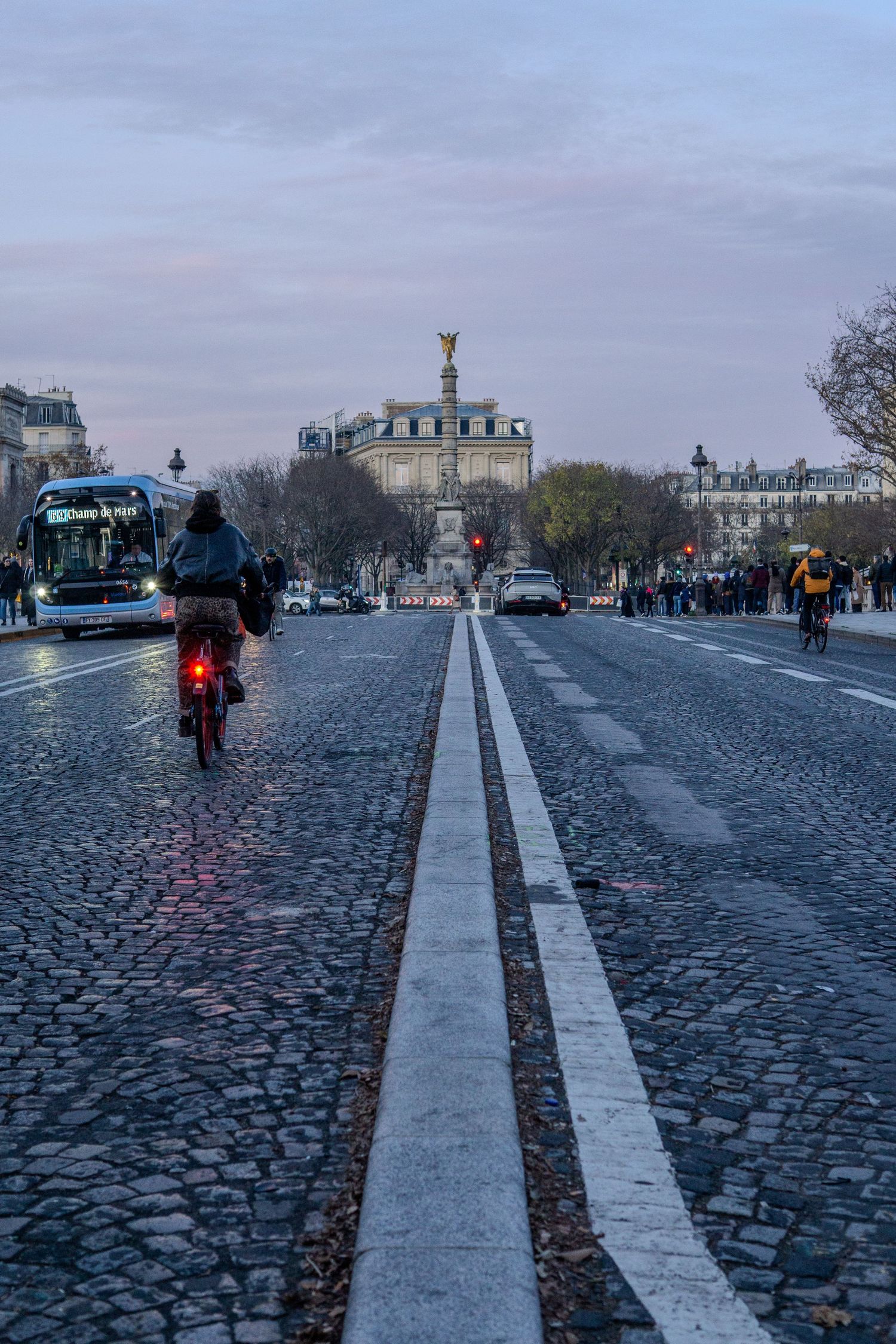Cobblestone street in Paris with cyclist and Bastille monument visible in distance at dusk.