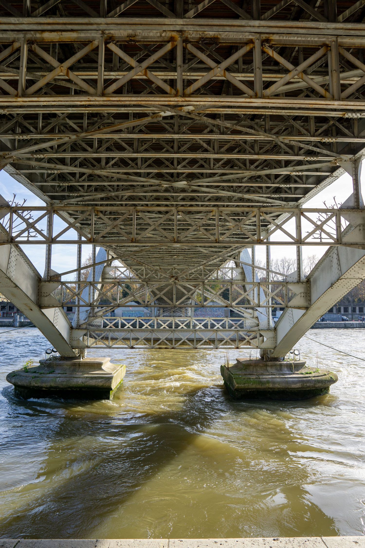 View underneath a steel bridge showing its support structure and pillars over muddy brown river water.