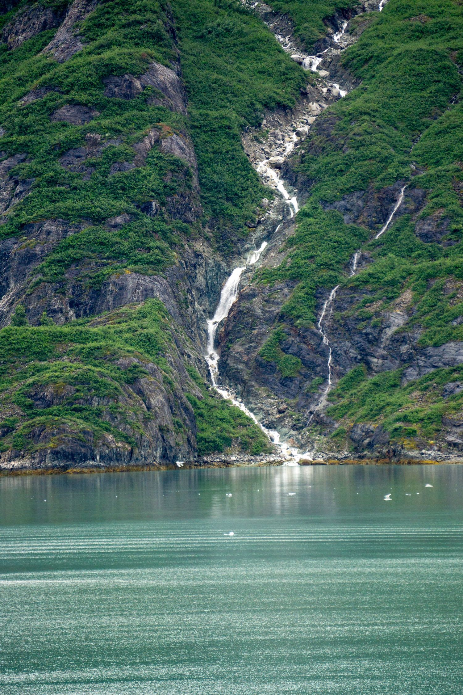 A dramatic waterfall cascades down a steep green mountainside into turquoise glacial waters in Alaska.
