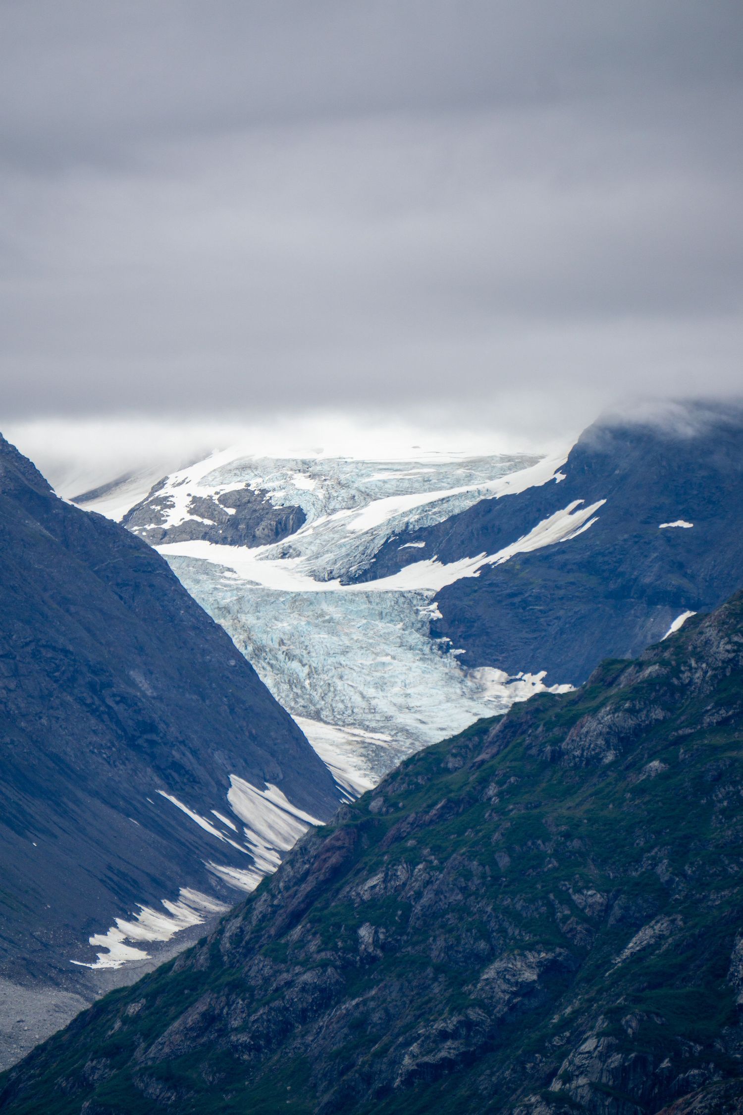 Ancient glacier carves its way through steep mountain valleys beneath overcast skies.