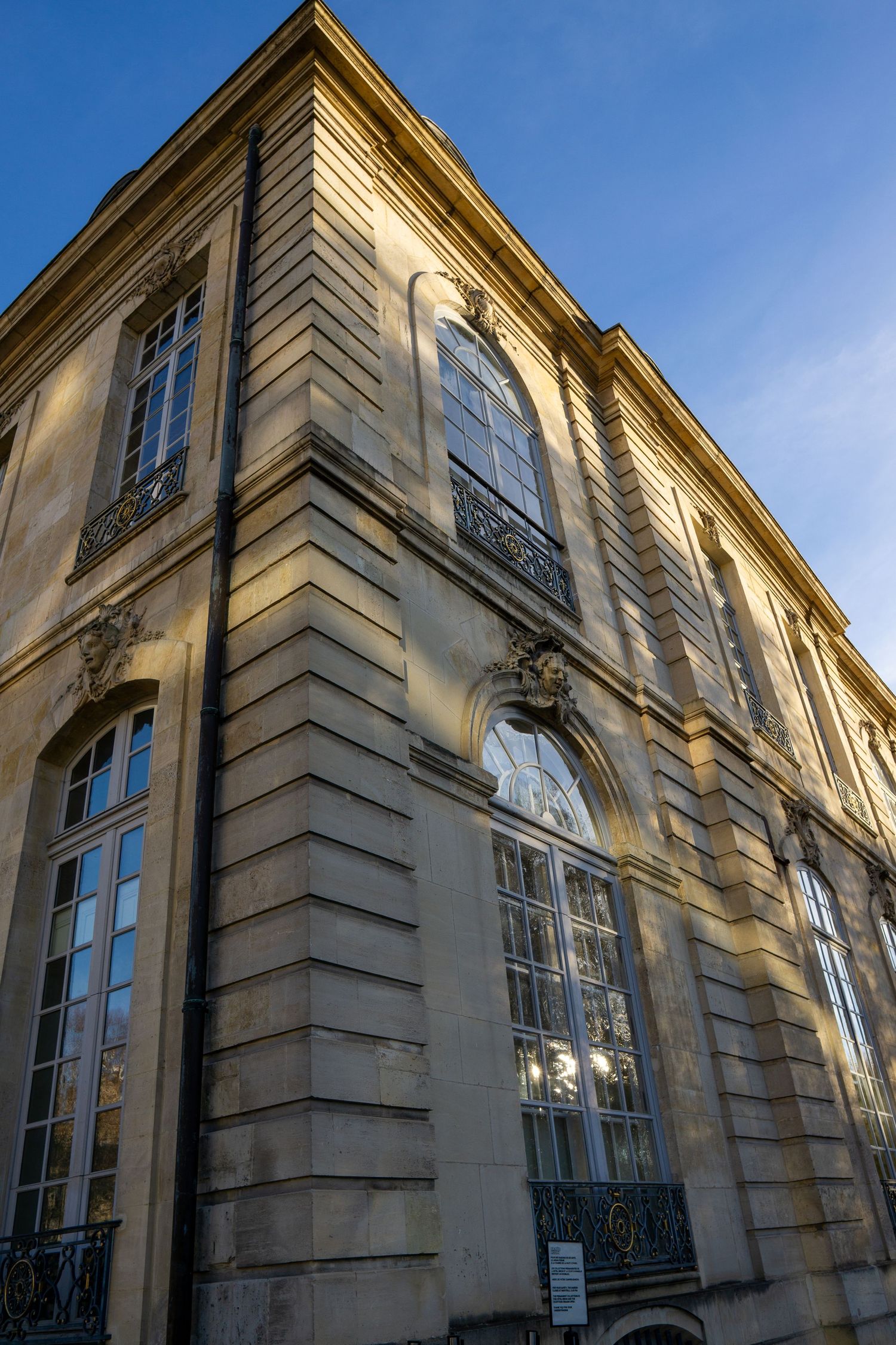 Classical French architecture with tall arched windows and ornate stonework facade illuminated by warm sunlight against blue sky.