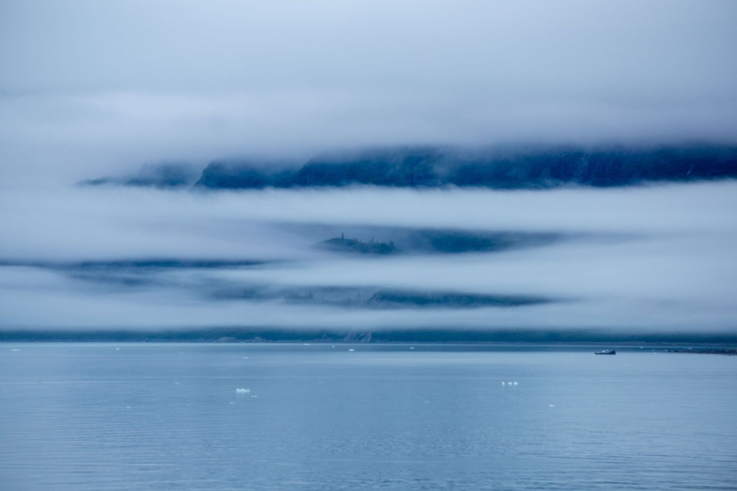 Ethereal fog layers hover over calm waters beneath a cloudy sky in Alaska.