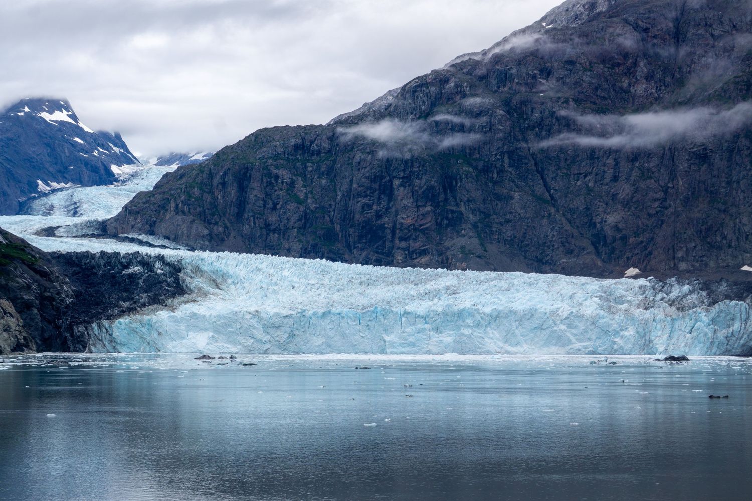 Majestic blue glacier terminus reflects in calm fjord waters beneath cloudy skies.