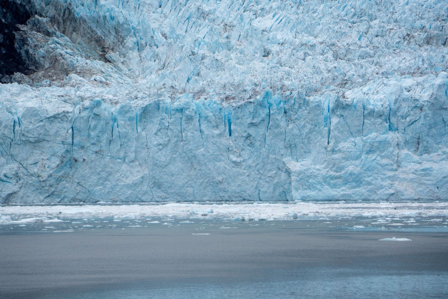 Textured blue ice wall of a massive glacier reflects in calm fjord waters.