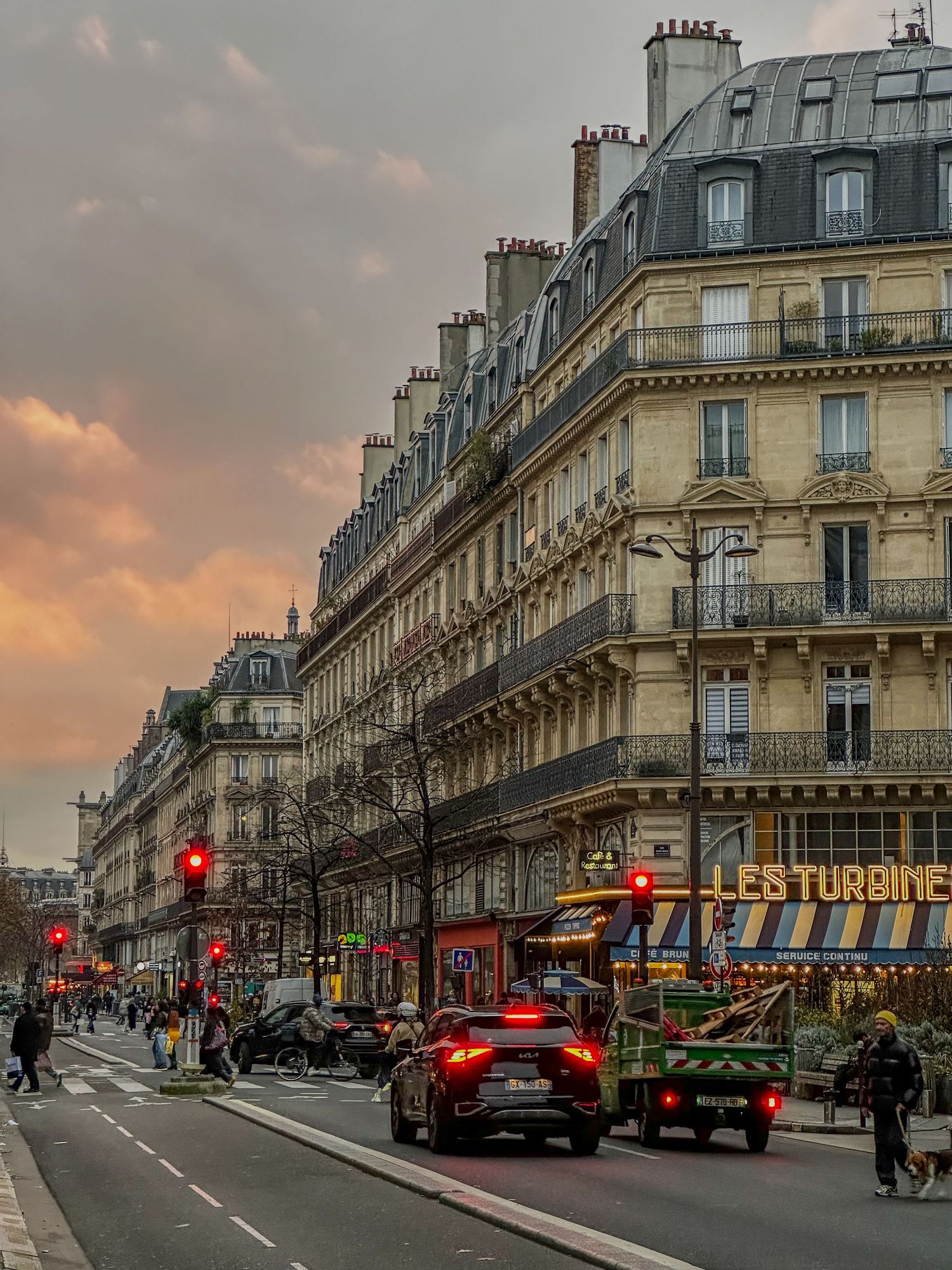 Classic Parisian street with Haussmann-style architecture at sunset, featuring traffic lights and storefronts on a cloudy evening.