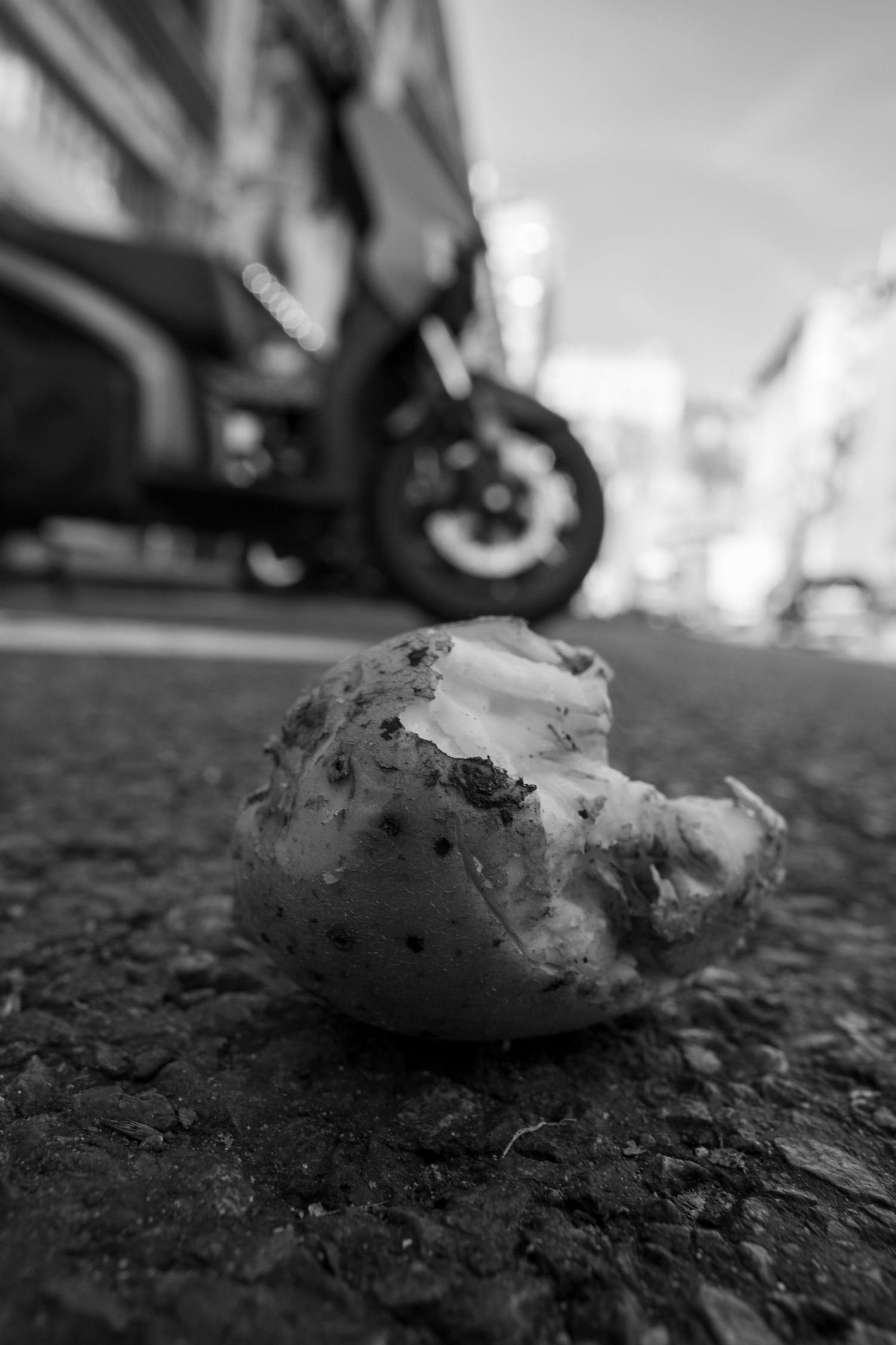 A bitten donut lies discarded on asphalt with a motorcycle blurred in the background, shot in black and white.