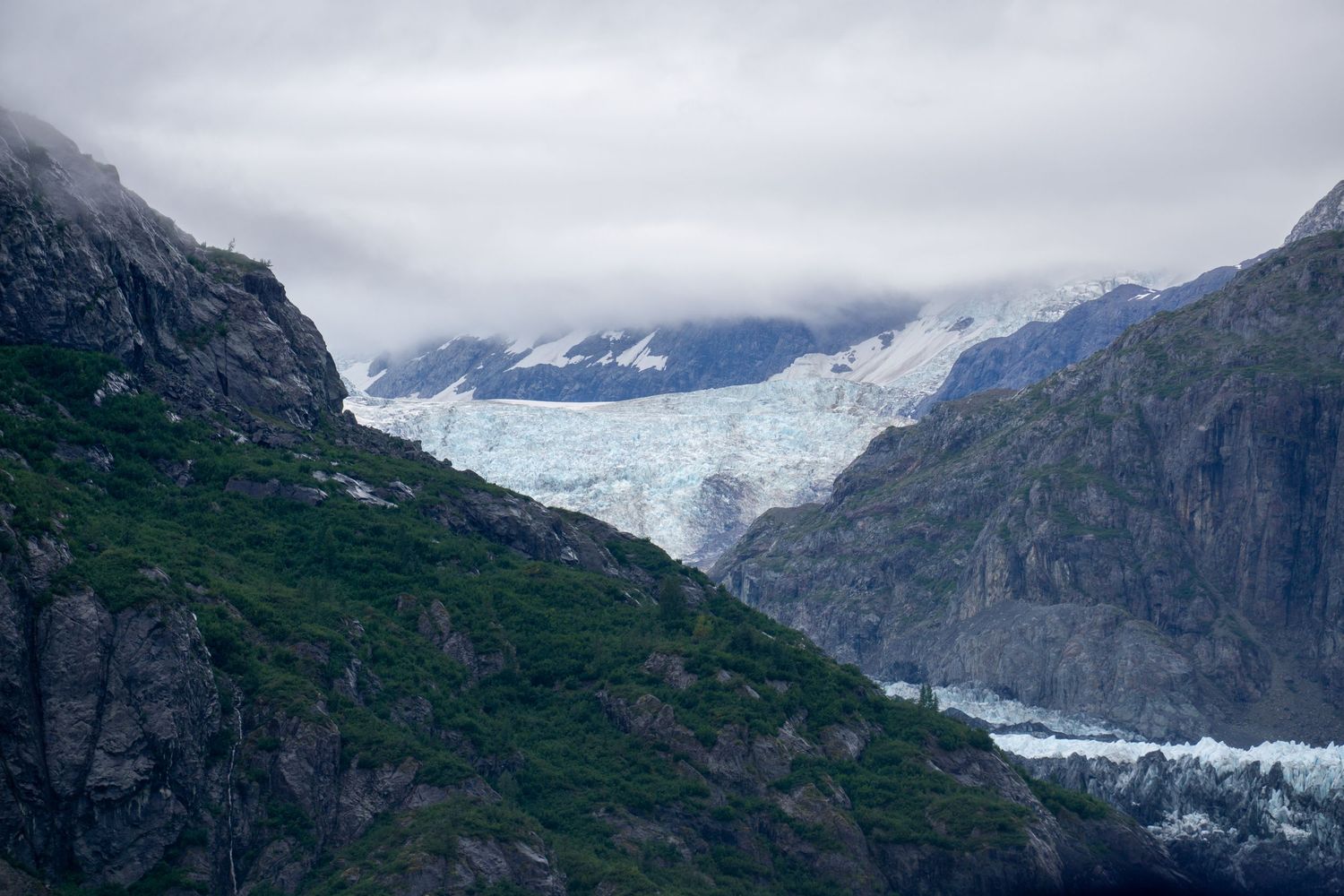 Glacier flows between rocky mountain peaks under cloudy skies.
