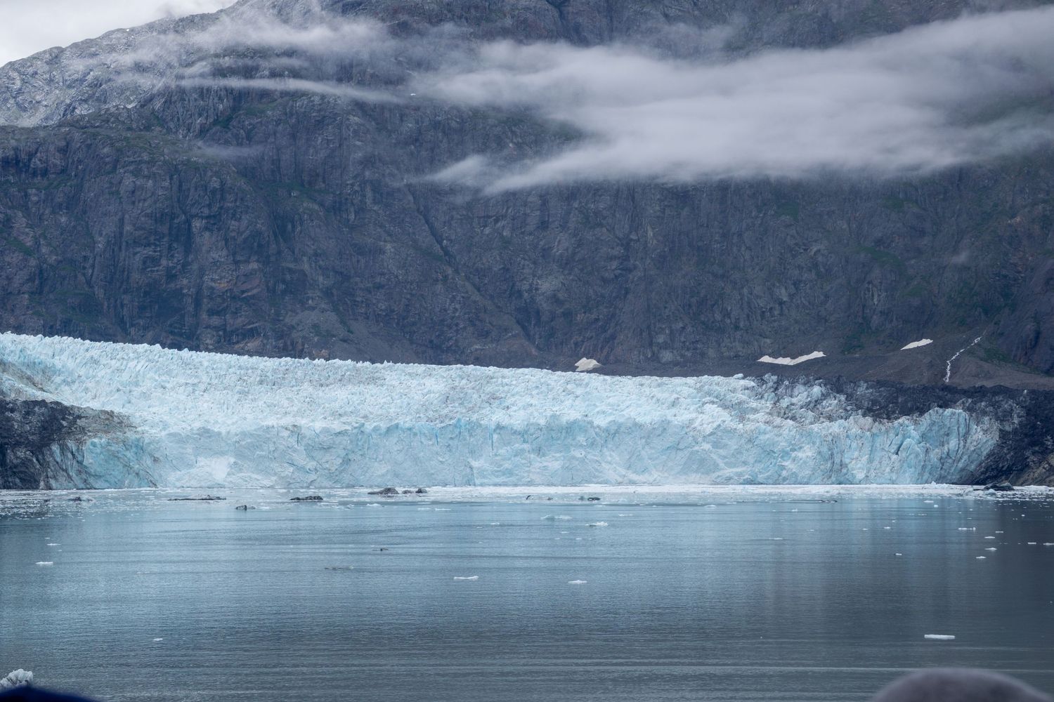 Massive glacier terminus meets calm waters beneath fog-shrouded mountain cliffs.