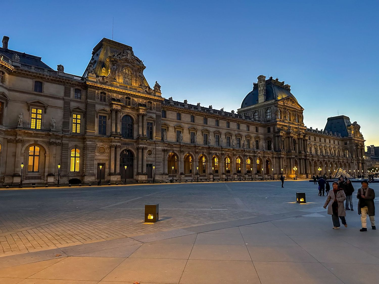 Evening view of the illuminated Louvre Museum facade in Paris with tourists walking along the courtyard plaza at dusk.