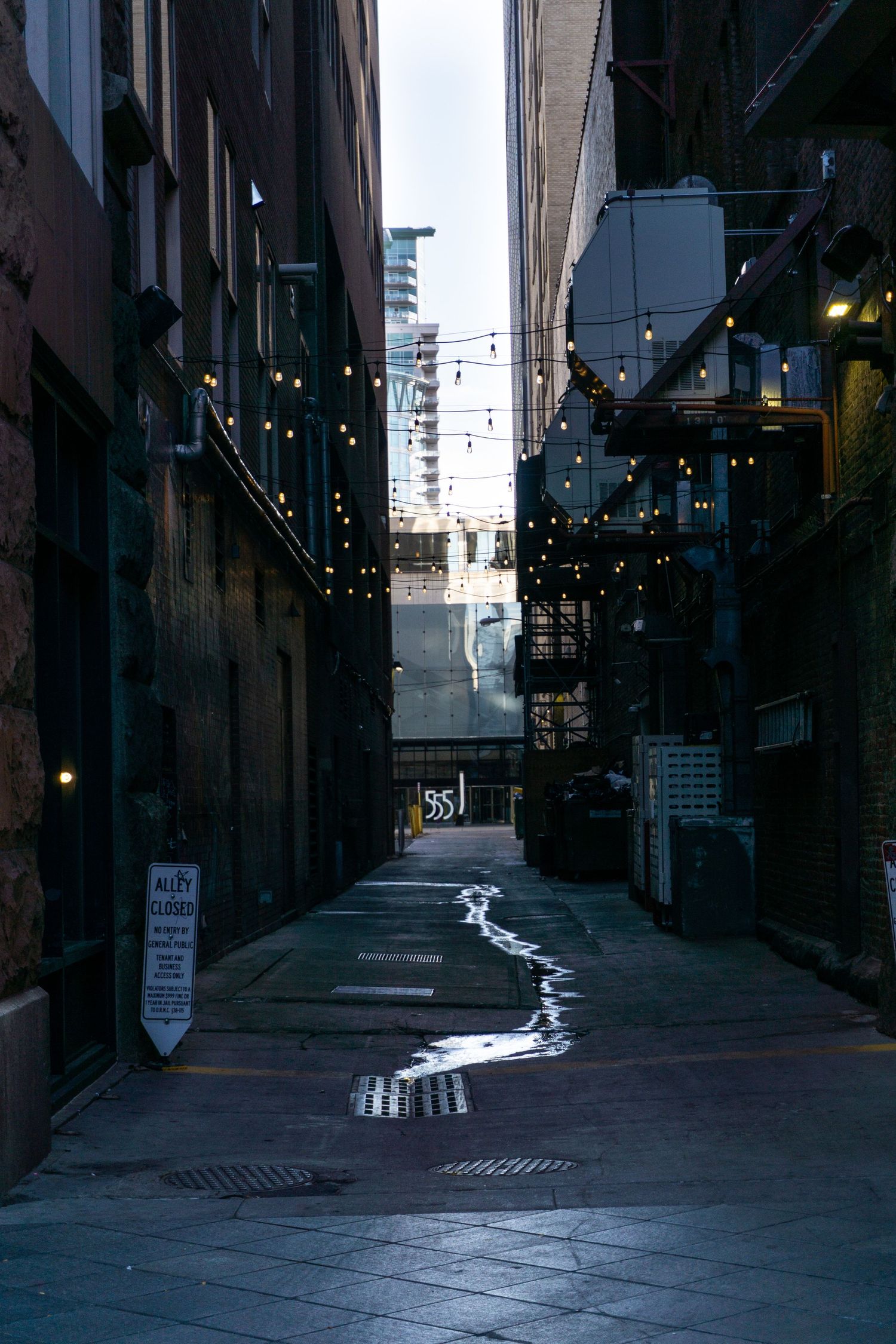 Urban alley with string lights and a winding water trail on cobblestone ground.