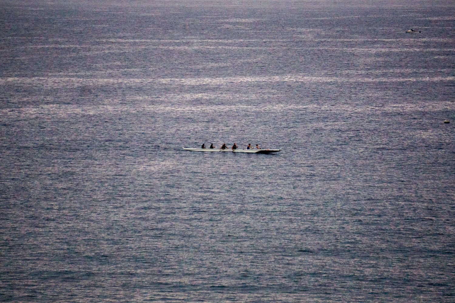 A distant rowing crew glides across the calm ocean waters at dusk.