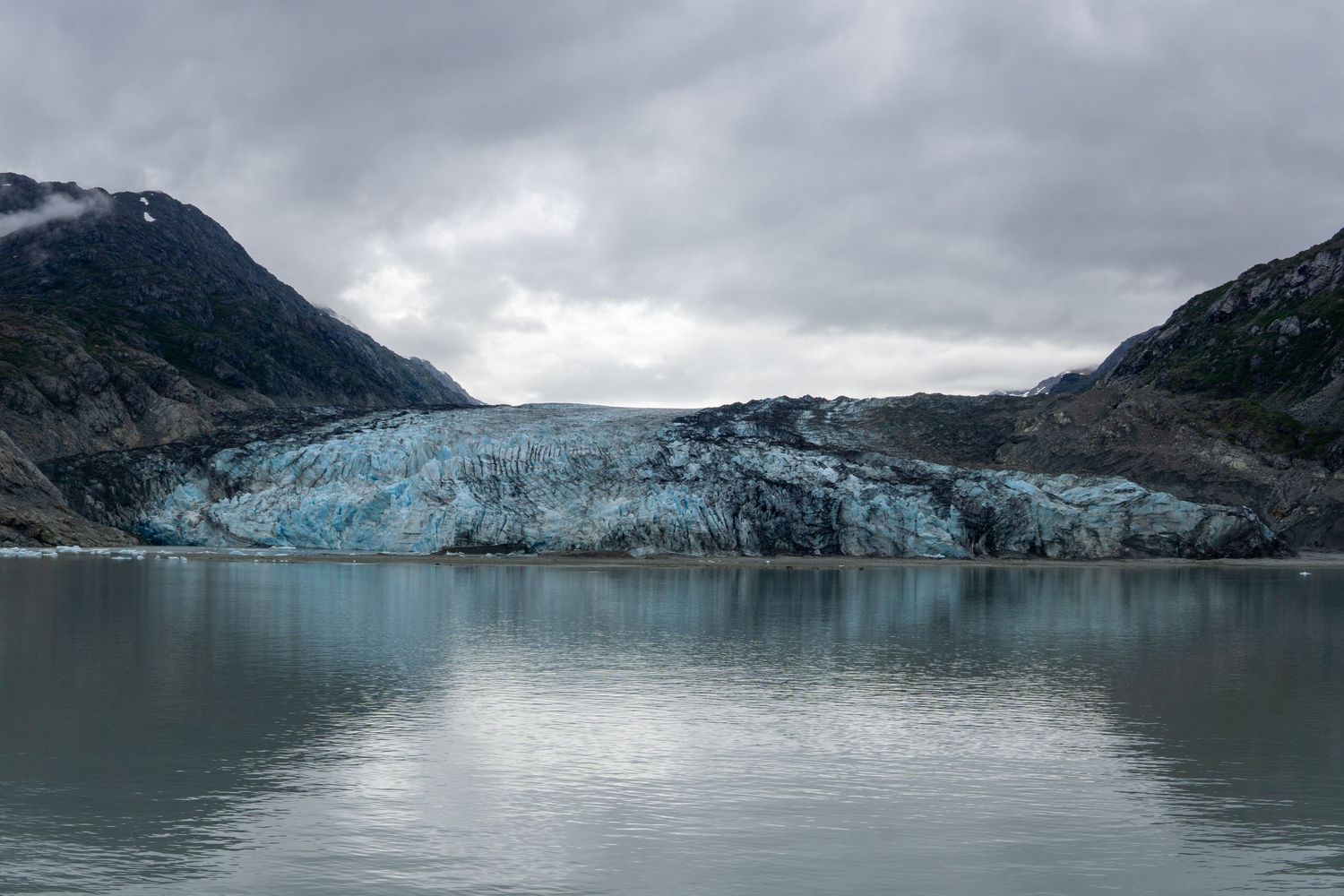 Blue glacier terminus stretches across mountain valley reflecting in still waters below overcast sky.