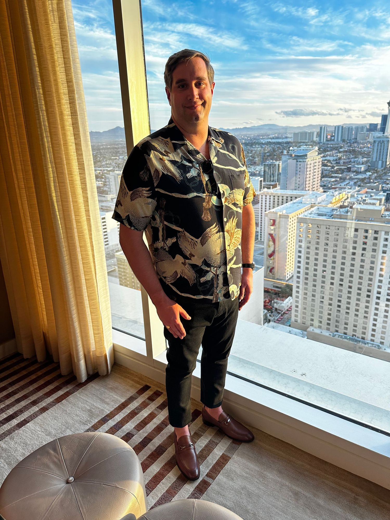 Person wearing floral shirt standing by high-rise hotel window overlooking the Las Vegas skyline.