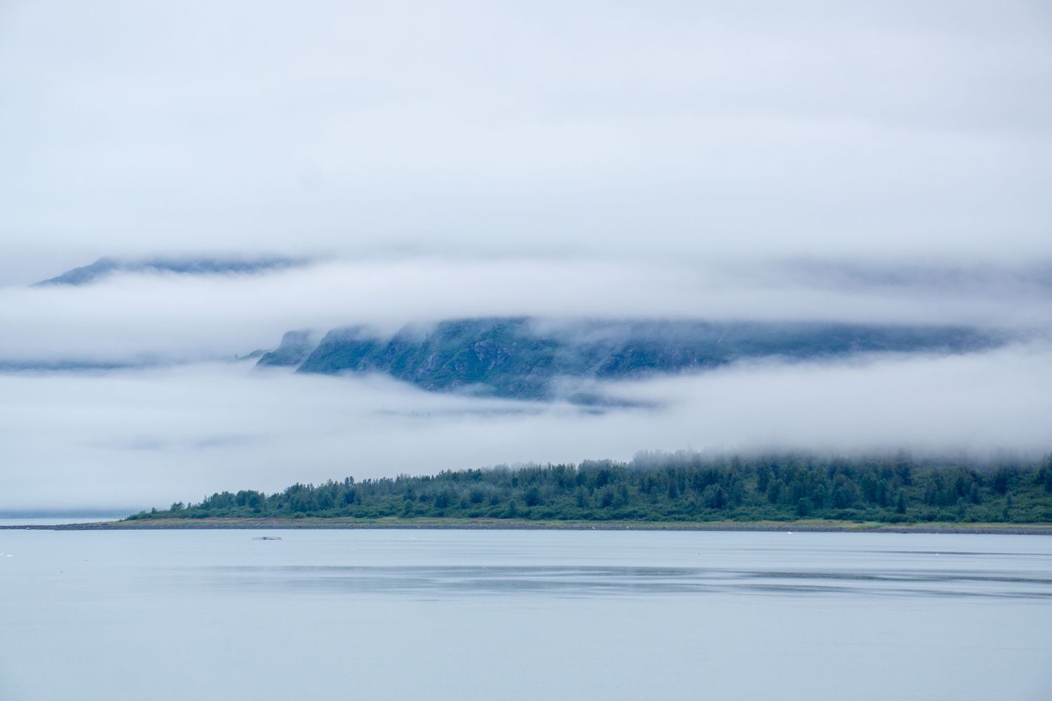 Layers of fog wrap around blue mountains reflected in still waters.