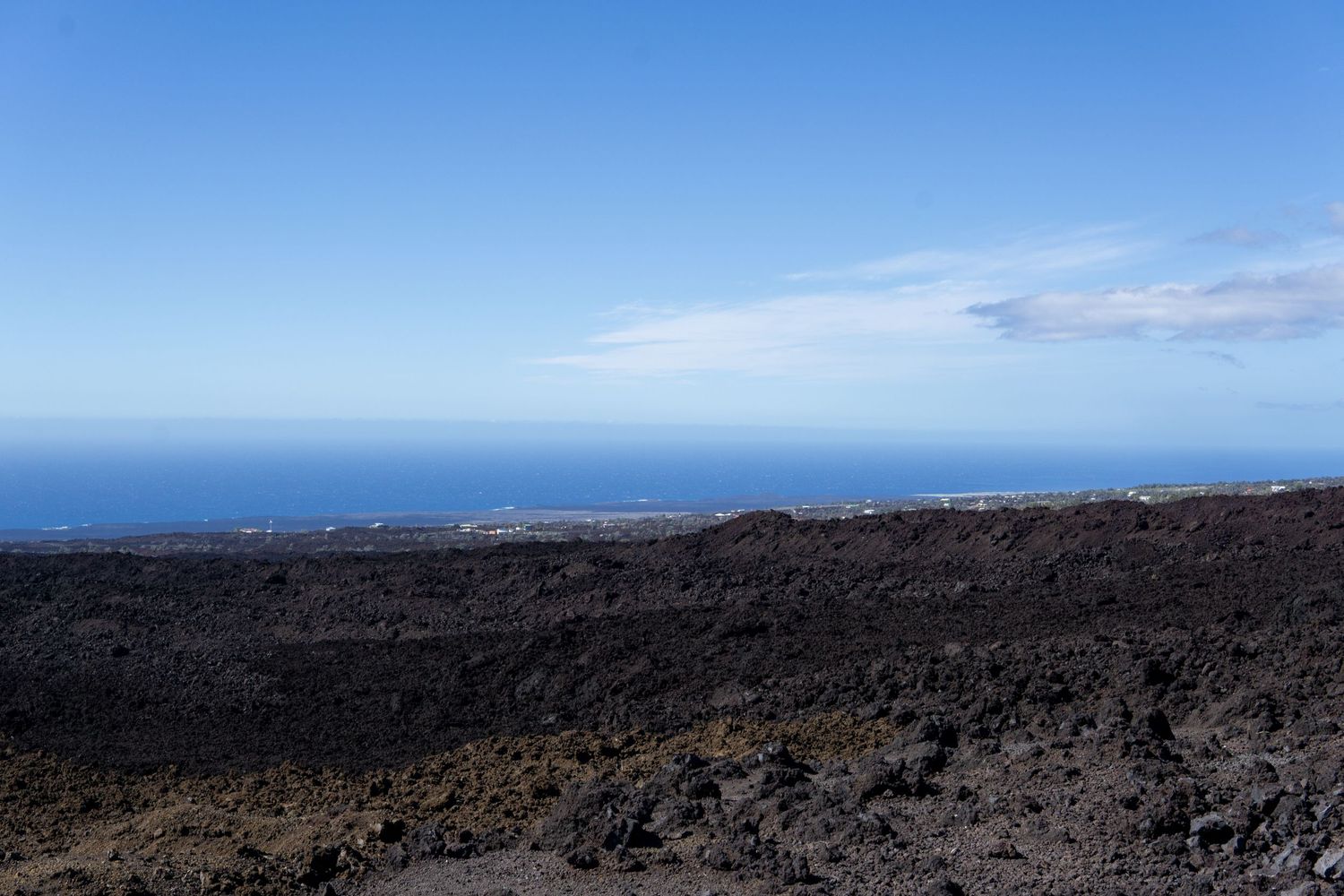 Volcanic landscape with black lava rocks stretches toward blue ocean horizon under clear Hawaiian sky.