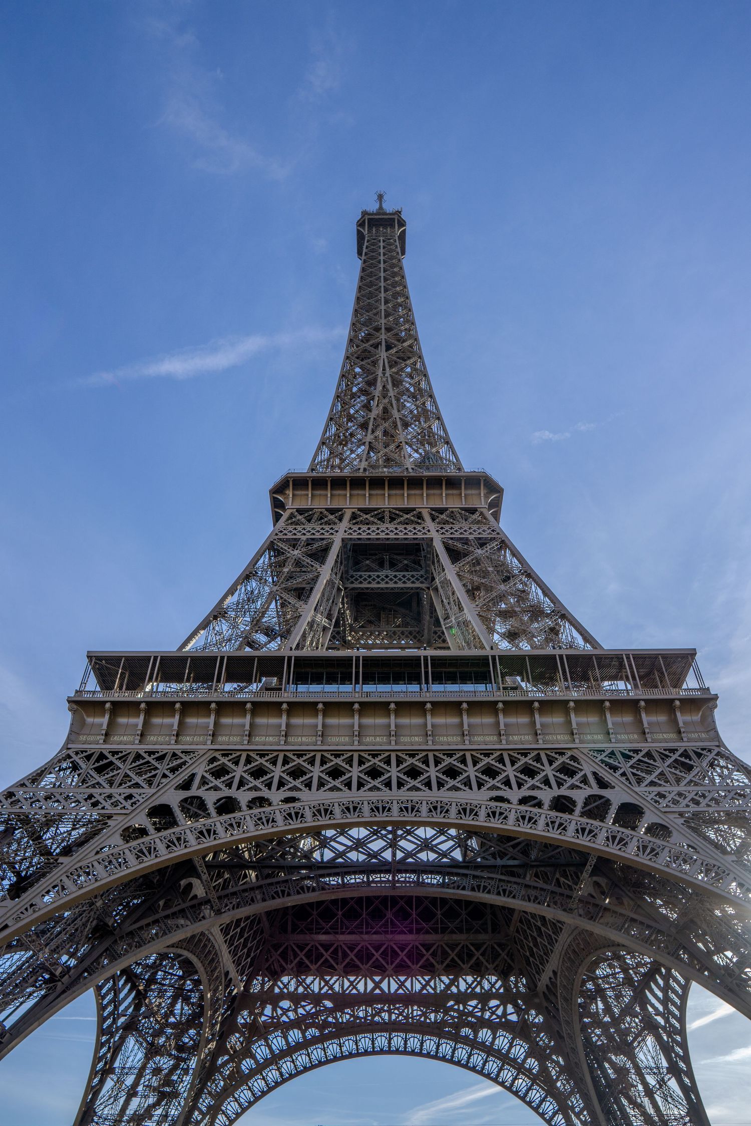 Looking up at the iconic Eiffel Tower in Paris against a blue sky, showcasing its intricate iron lattice architecture.