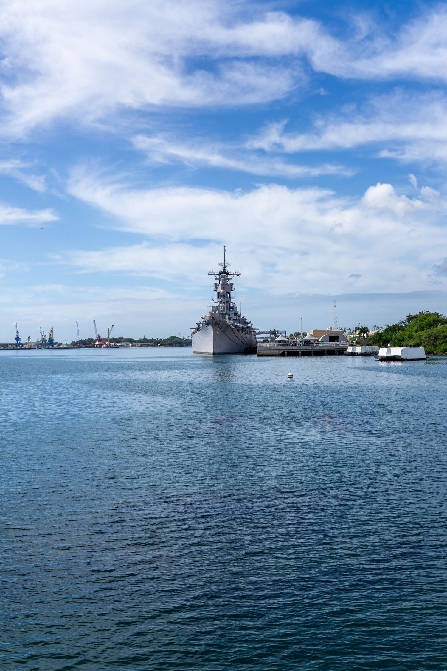 USS Missouri battleship docked in Pearl Harbor with blue skies and scattered clouds above the calm waters.