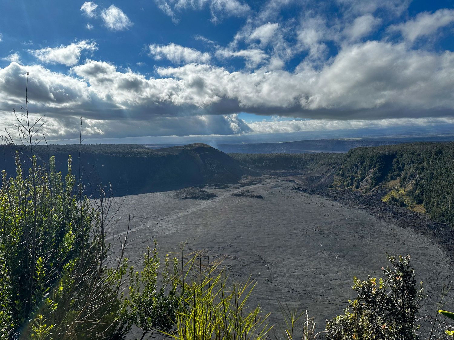 Aerial view of Kilauea Iki crater in Hawaii Volcanoes National Park with blue sky and puffy clouds above.