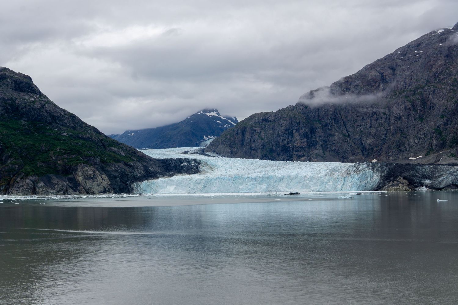Blue glacier face stretches across a fjord between steep mountain walls under overcast skies.