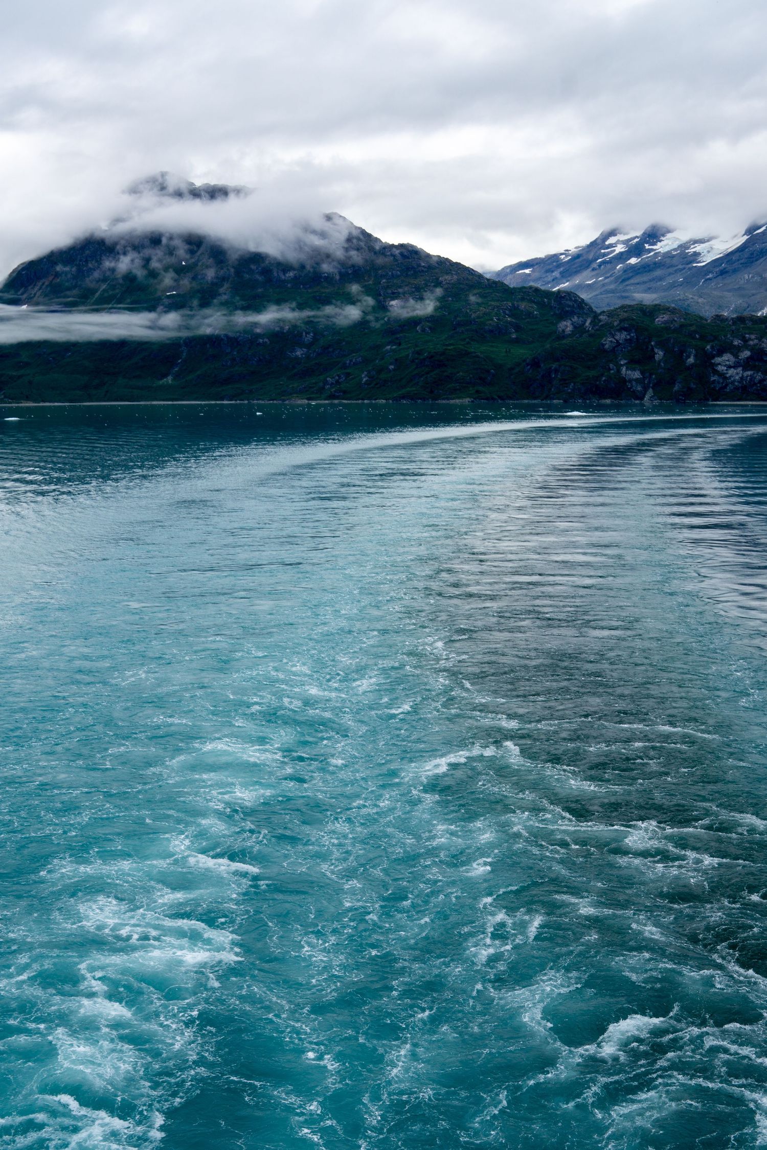 Ship's wake creates patterns in turquoise glacial waters beneath misty mountain peaks.