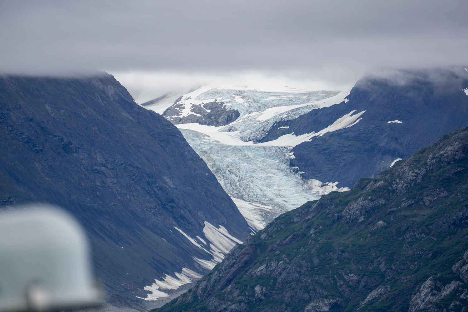 Massive glacier flows between mountain peaks under cloudy skies in Alaska.