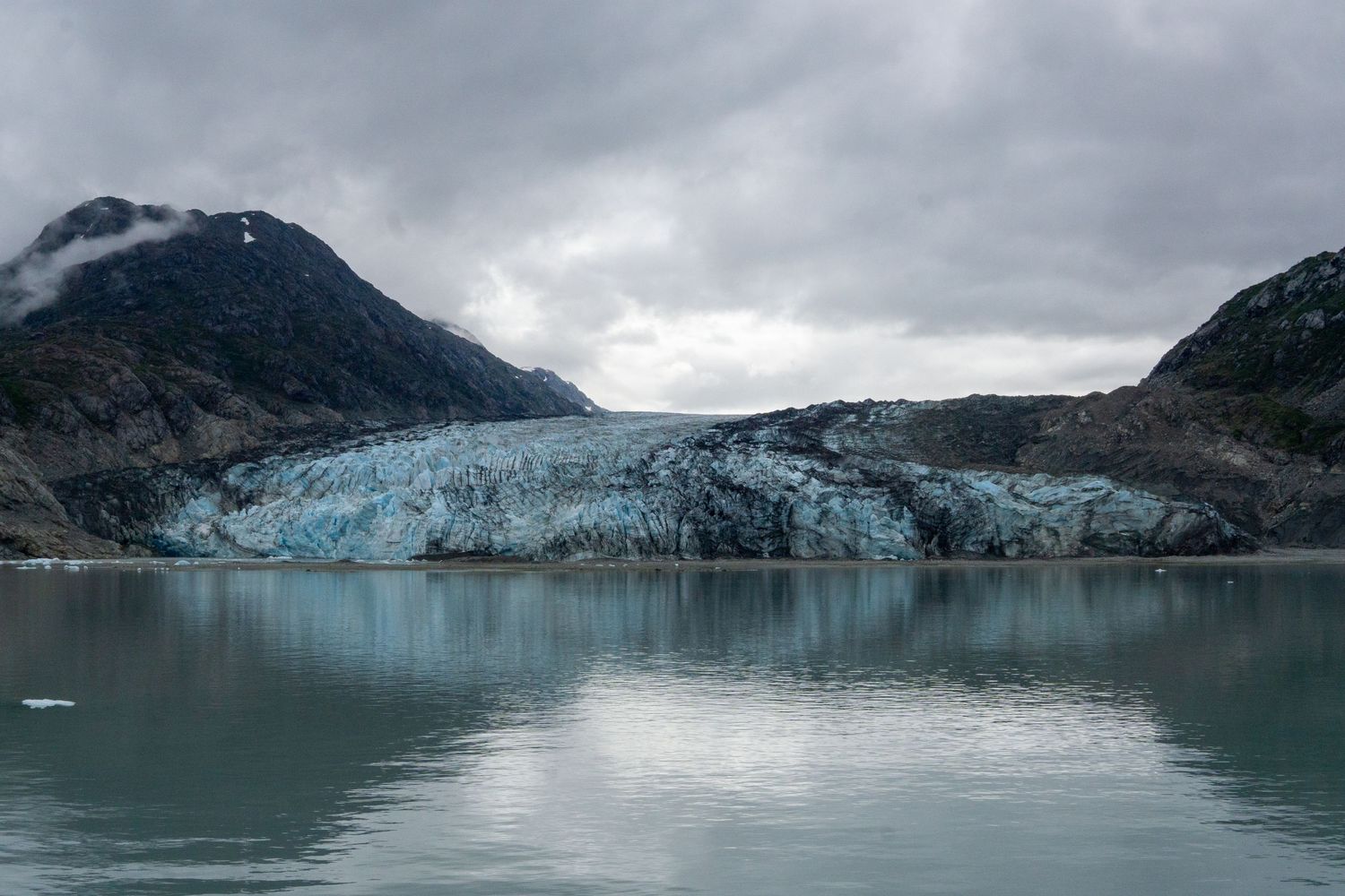 Massive glacier with blue ice meets turquoise waters under dramatic cloudy skies in a fjord landscape.