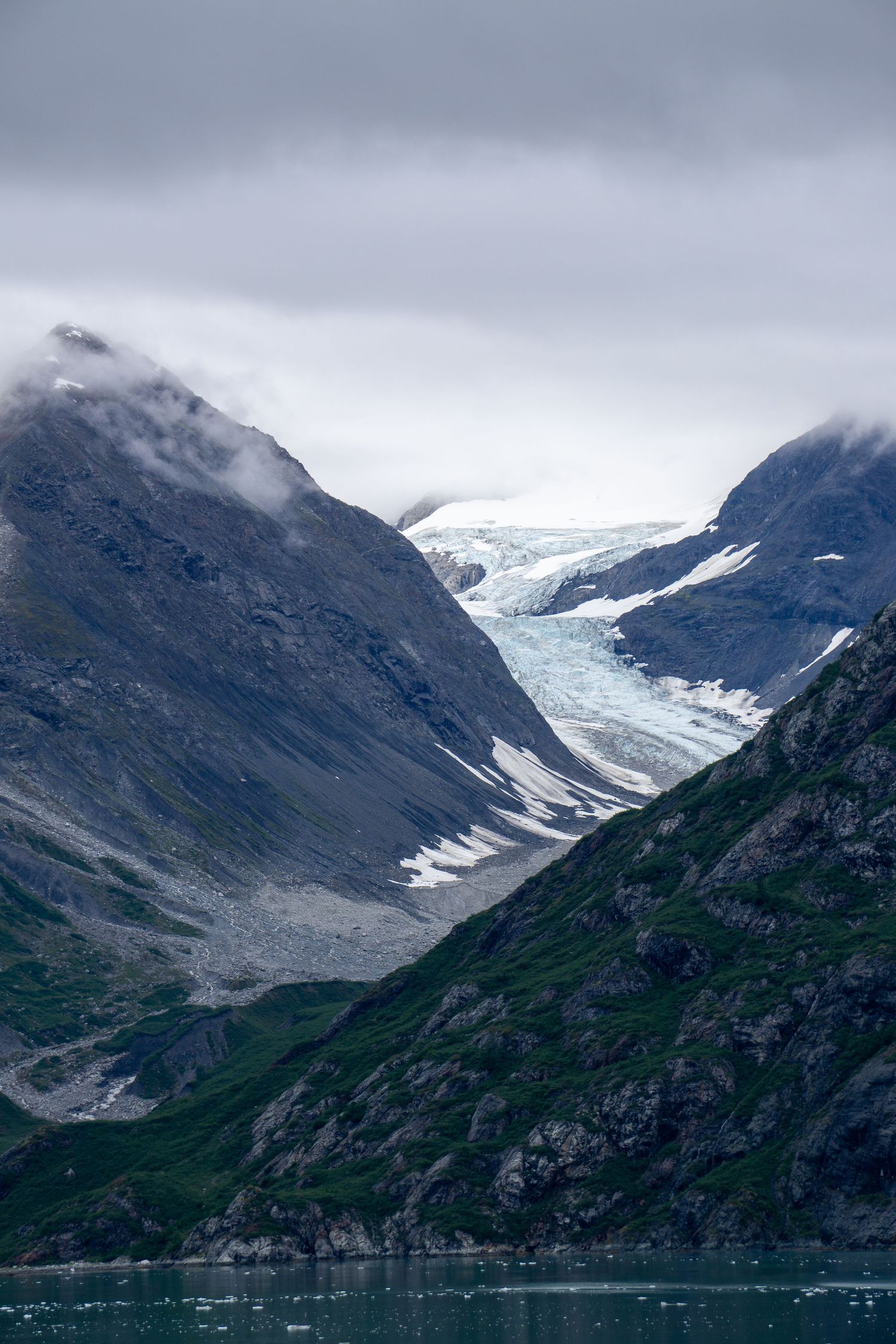 Massive glacier descends through steep mountain valley toward calm fjord waters.