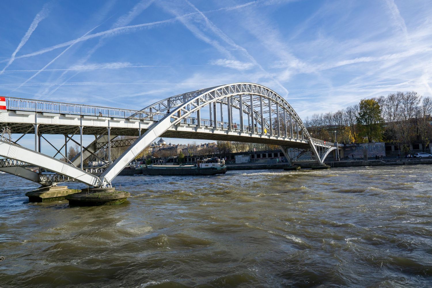 White arched steel bridge spans over rushing river waters under blue sky with contrail patterns in Paris.