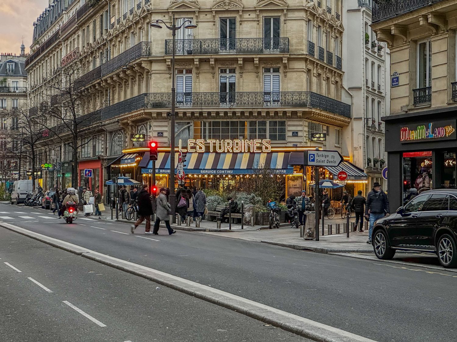 A lively street corner cafe in Paris with outdoor seating and typical Haussmann architecture in the evening light.