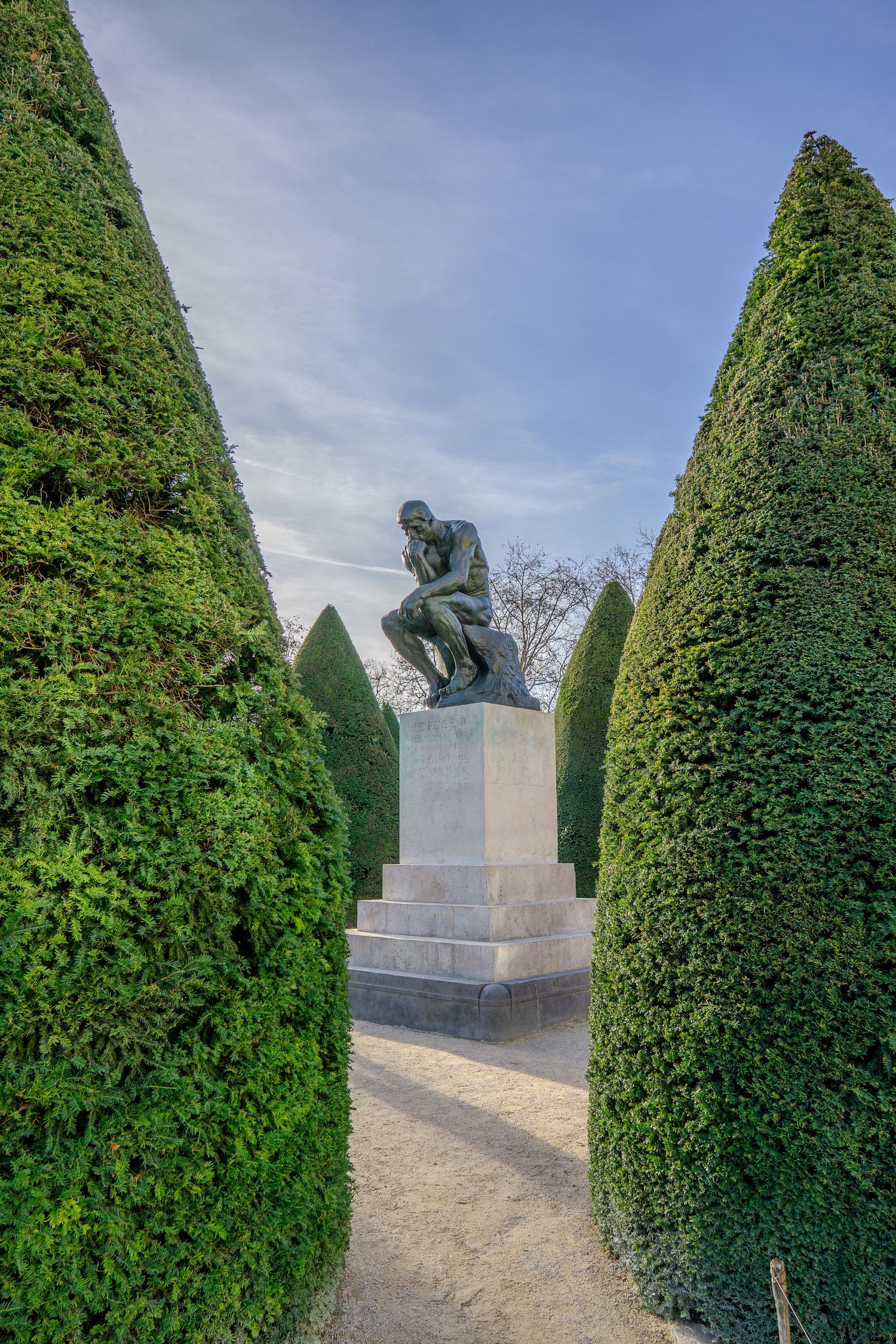 Rodin's famous Thinker statue framed by tall manicured hedges in a formal garden setting against a blue sky.