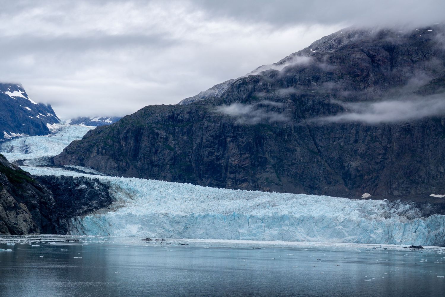 Massive blue glacier terminus spans between mountains in an Alaskan fjord.