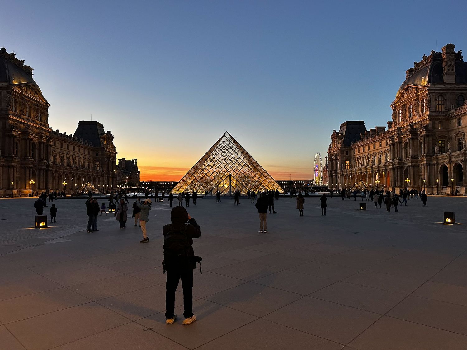 Louvre Museum pyramid glows at sunset with visitors silhouetted against the iconic glass structure in Paris.