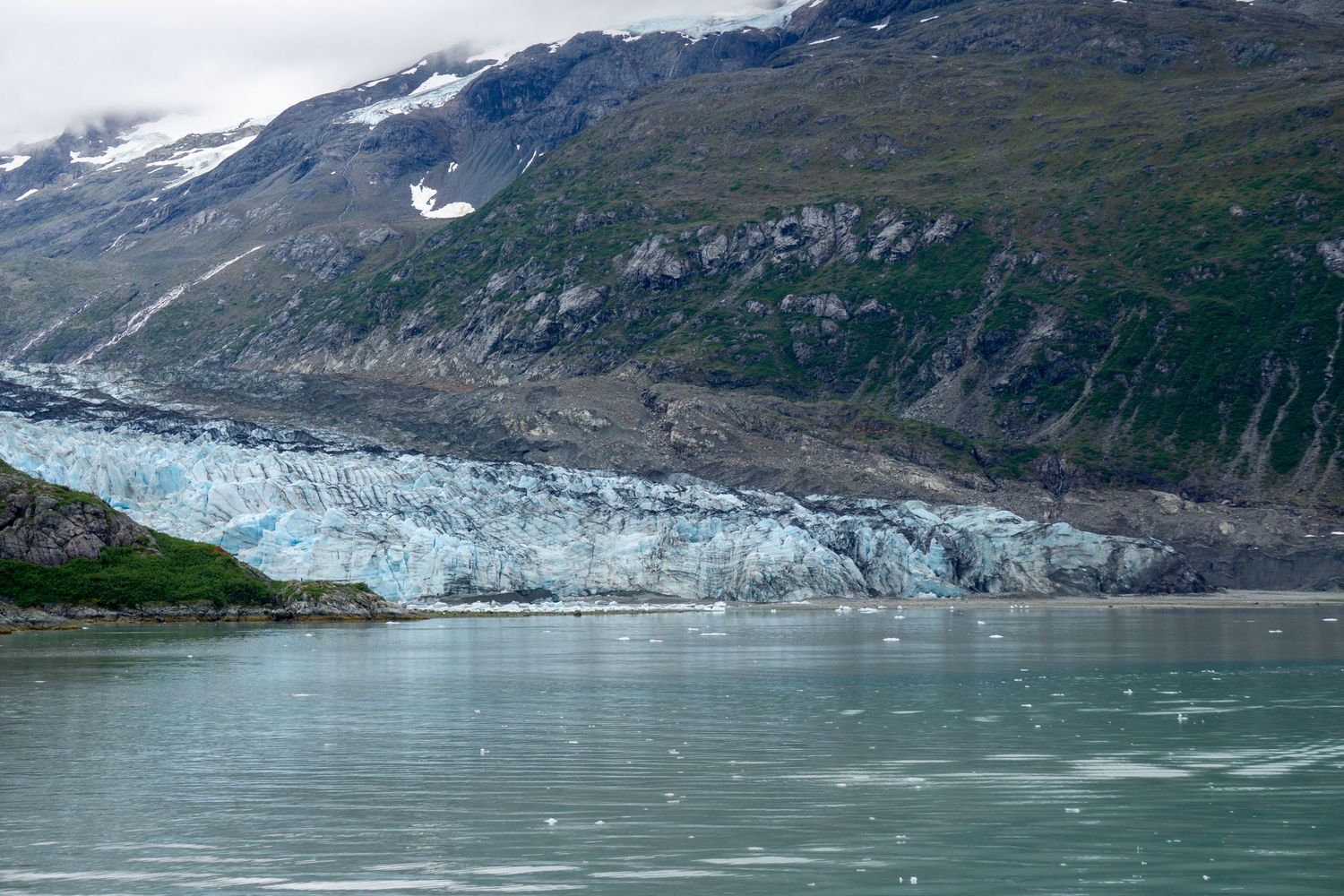 Massive glacier terminus meets calm waters surrounded by rugged mountain terrain under cloudy skies.
