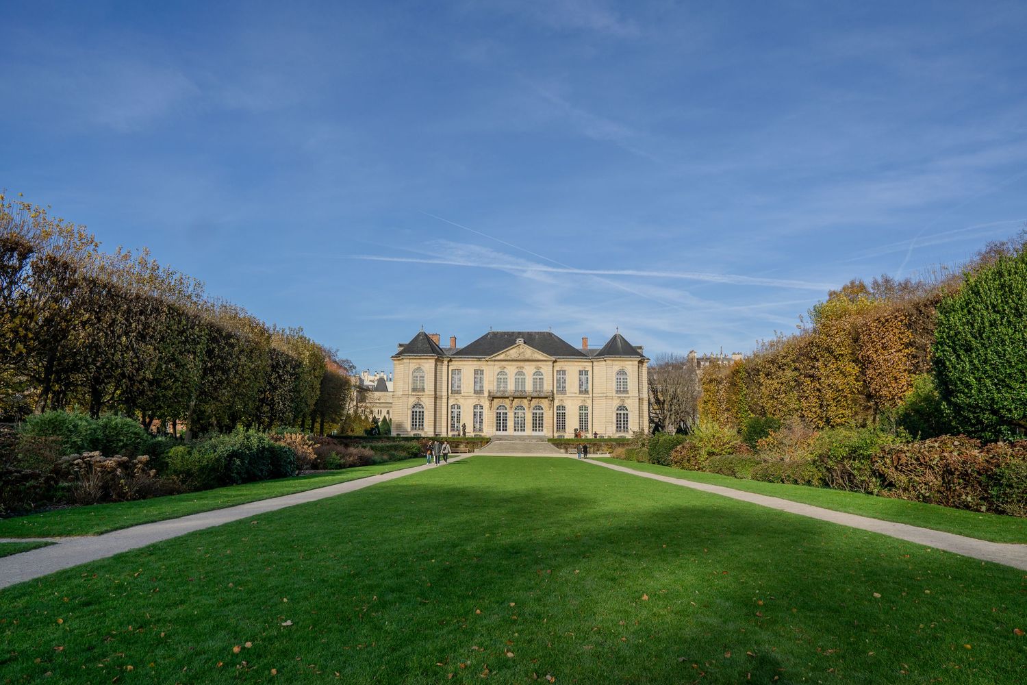 Grand mansion with symmetrical architecture set on manicured green lawn surrounded by autumn trees against blue sky.