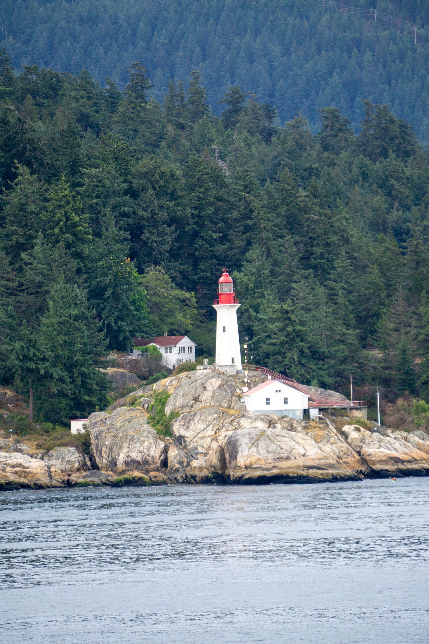 A white and red lighthouse perched on rocky cliffs surrounded by evergreen forest along a scenic coastline.