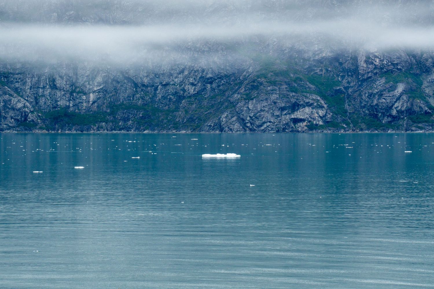 Turquoise glacial waters reflect rocky cliffs under misty skies with small icebergs floating.