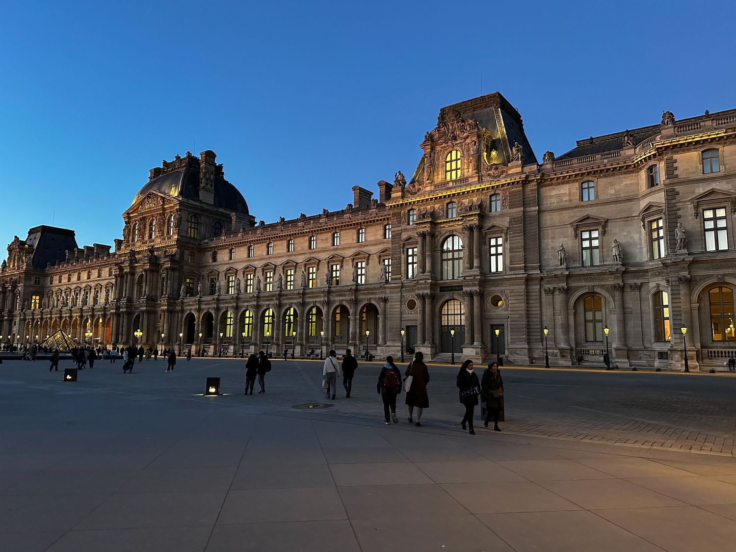 Exterior view of the Louvre Museum's ornate facade at dusk with people walking in front of the historic building in Paris.