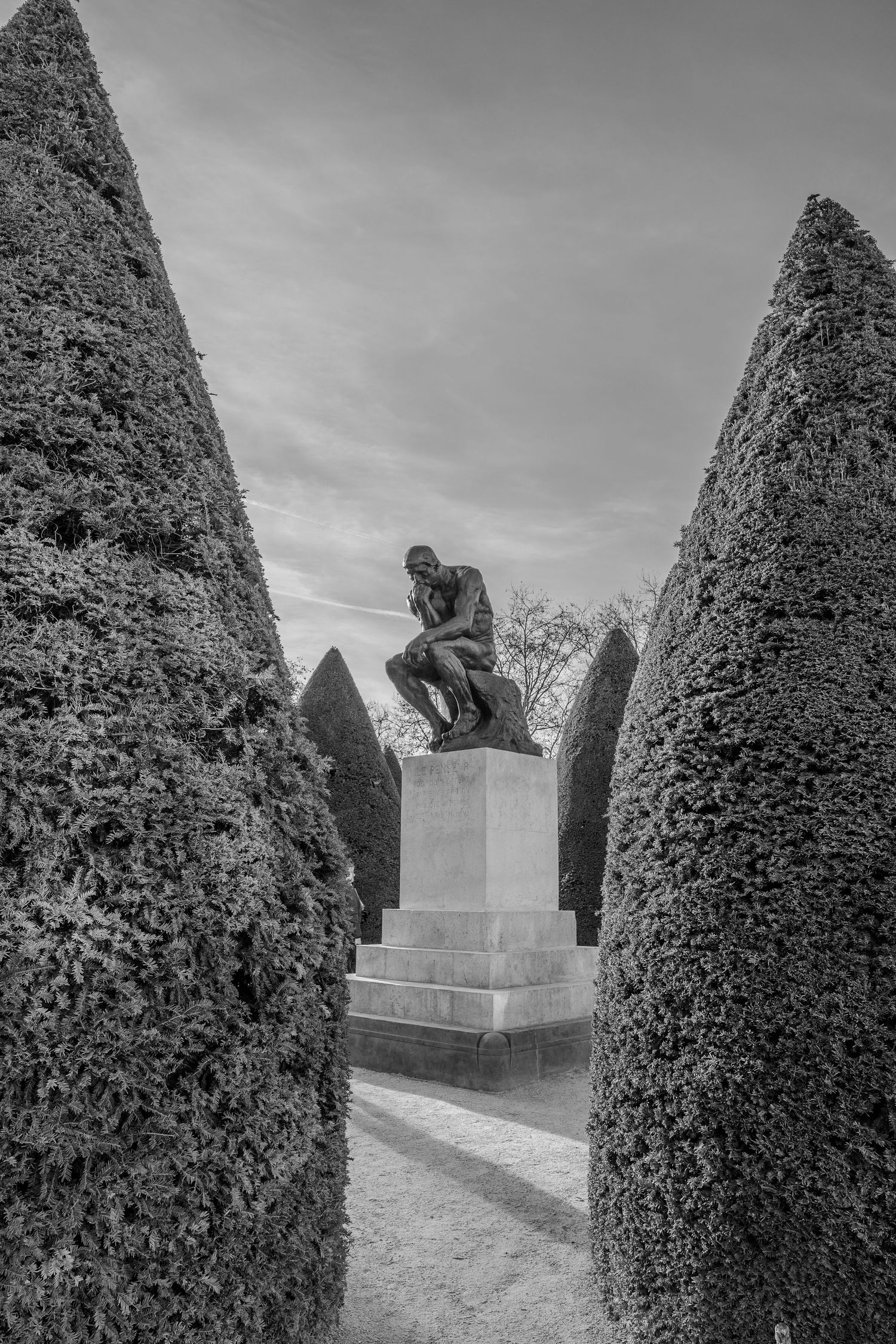 A bronze statue sits atop a pedestal between tall manicured hedges in a formal garden under cloudy skies.