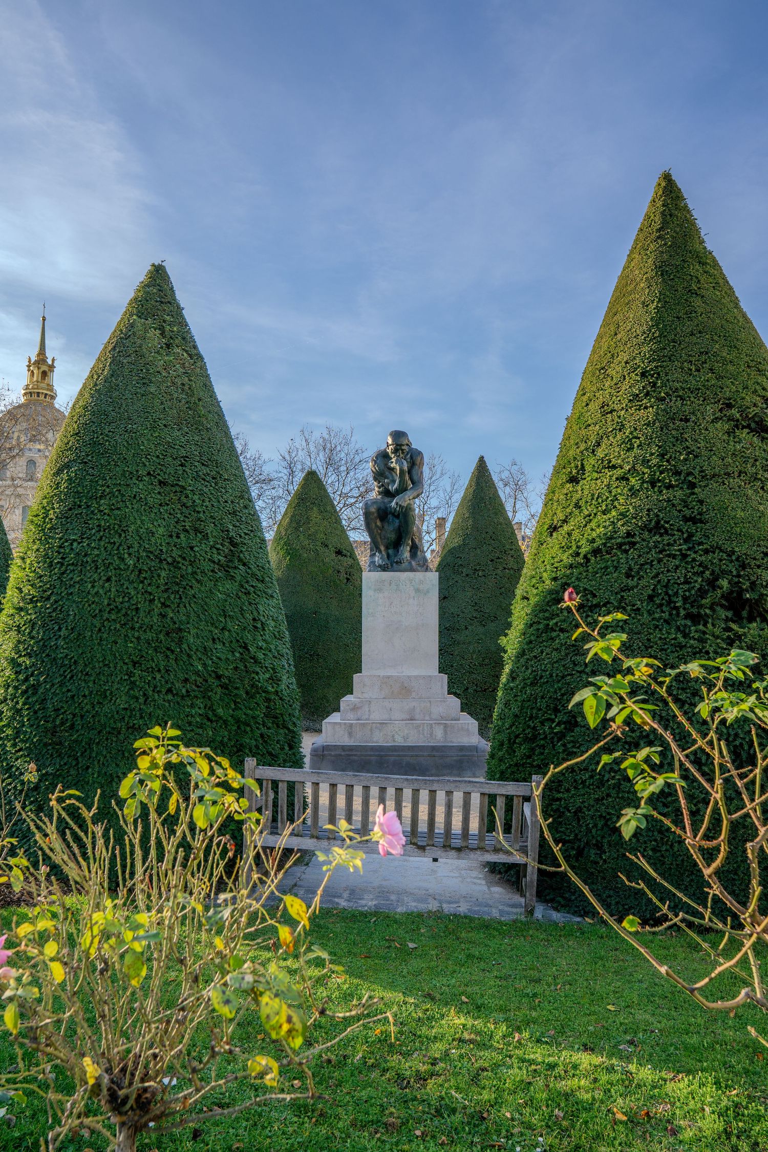 Bronze statue on pedestal surrounded by manicured conical evergreen trees and yellow flowers in a formal garden setting.