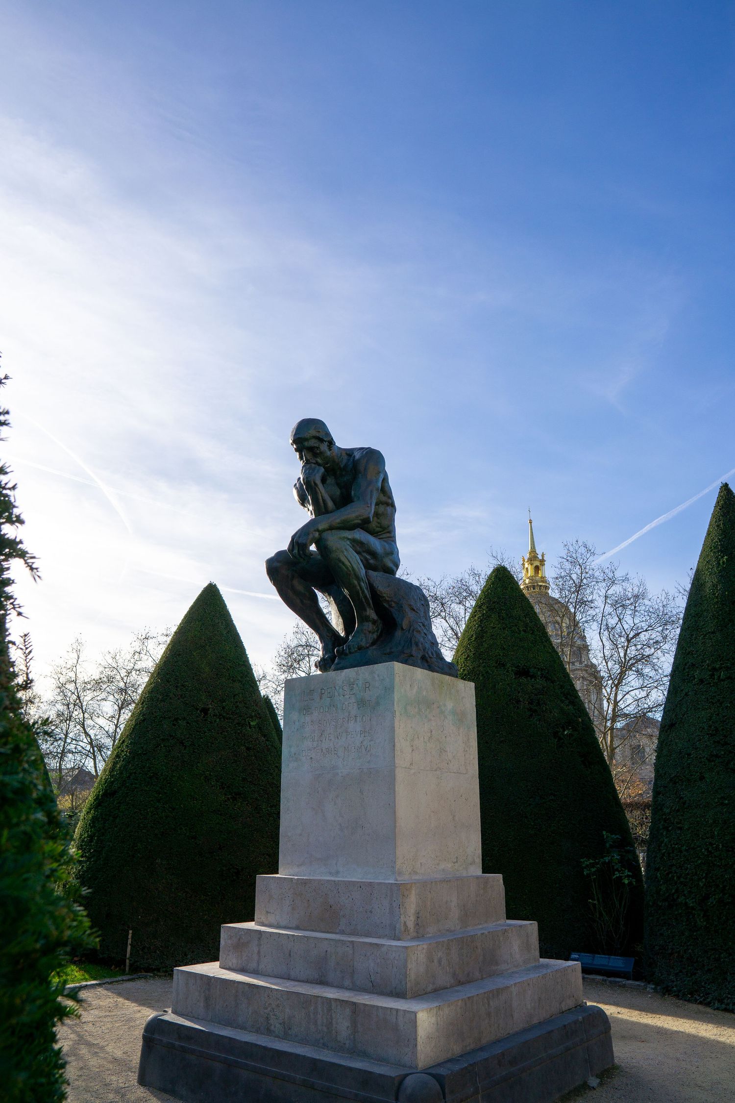 Rodin's The Thinker bronze sculpture sits atop stone pedestal surrounded by manicured topiary bushes under blue sky.
