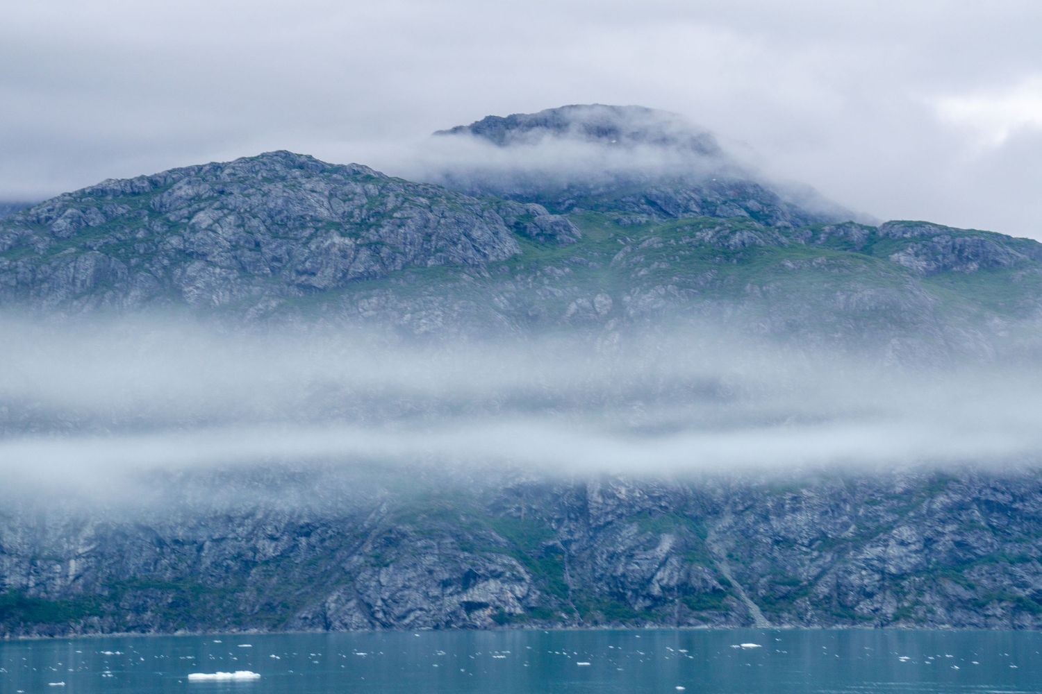 Rugged mountain peak draped in low clouds rising above glacial waters.